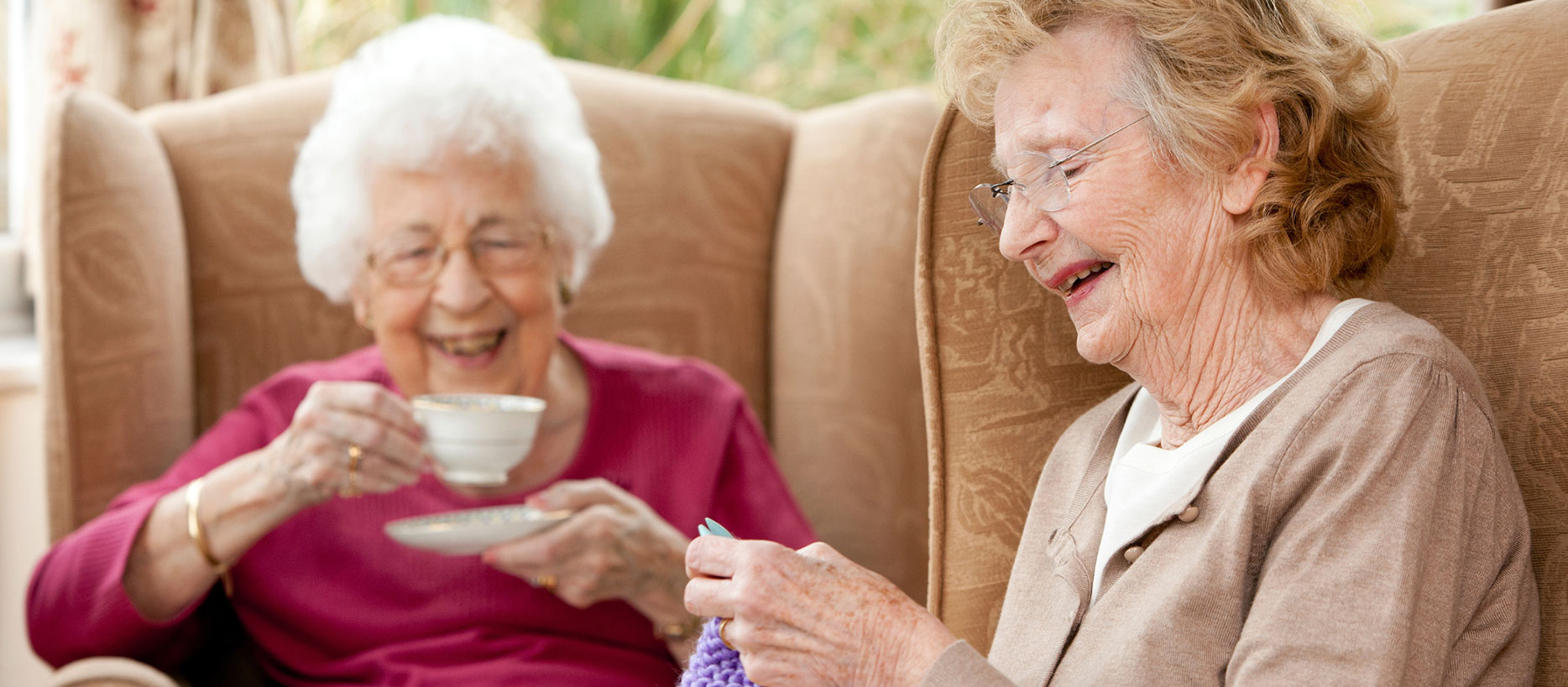 Two elderly women laughing, one drinking tea and one knitting